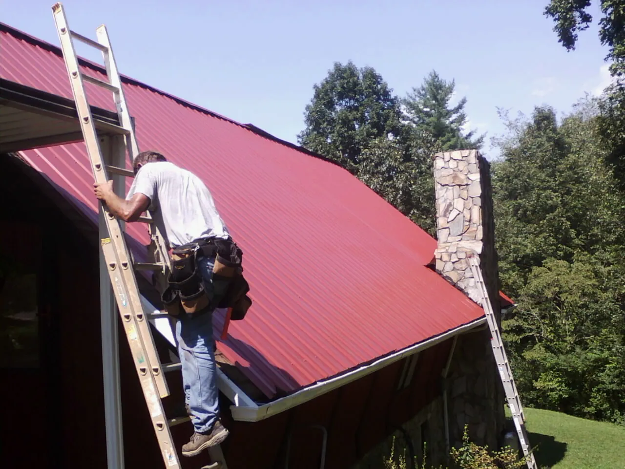 Roofer on a ladder beside a red metal roof and stone chimney — Soffit & Fascia Repair by Roofing Brothers in Musselshell